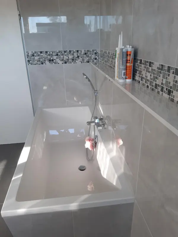 Badewanne mit Wannenarmatur vor grauen Fliesen mit Mosaikbordüre